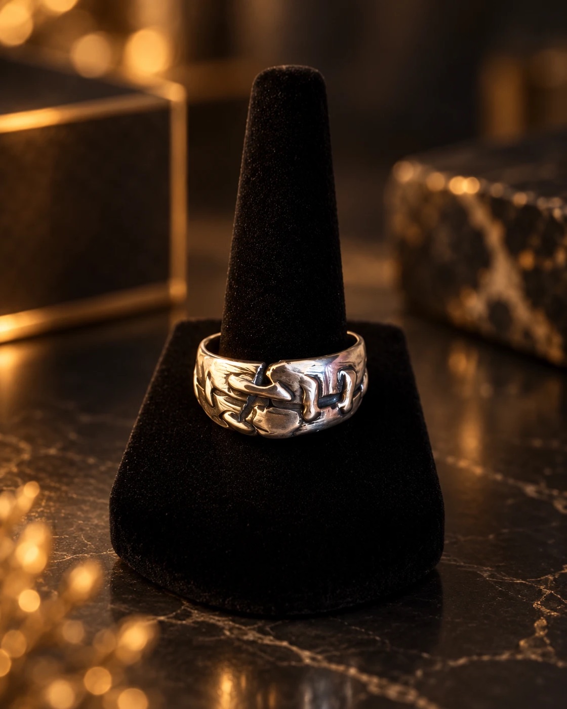Wide silver band sculpted with an interlocking stitched pattern, on a black velvet ring stand with warm bokeh behind